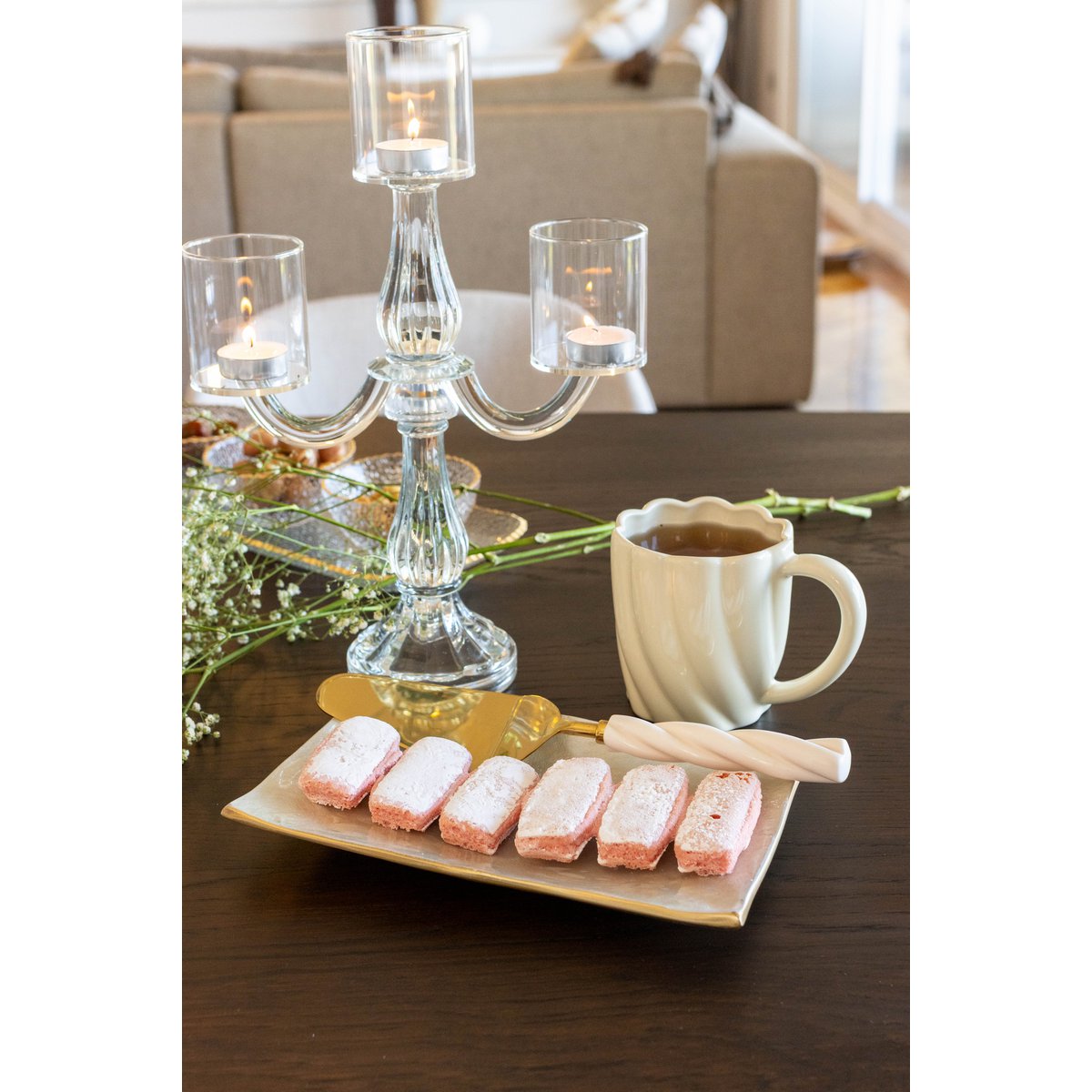 Clear glass candelabra with lit candles on a table with a cup of coffee and cookies.