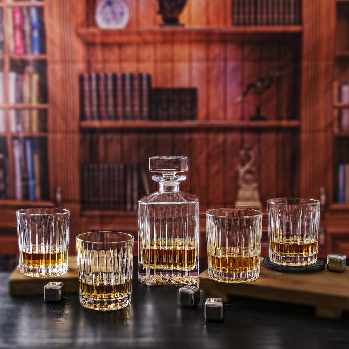 Set of whiskey glasses and a decanter on a wooden surface with a bookshelf in the background.